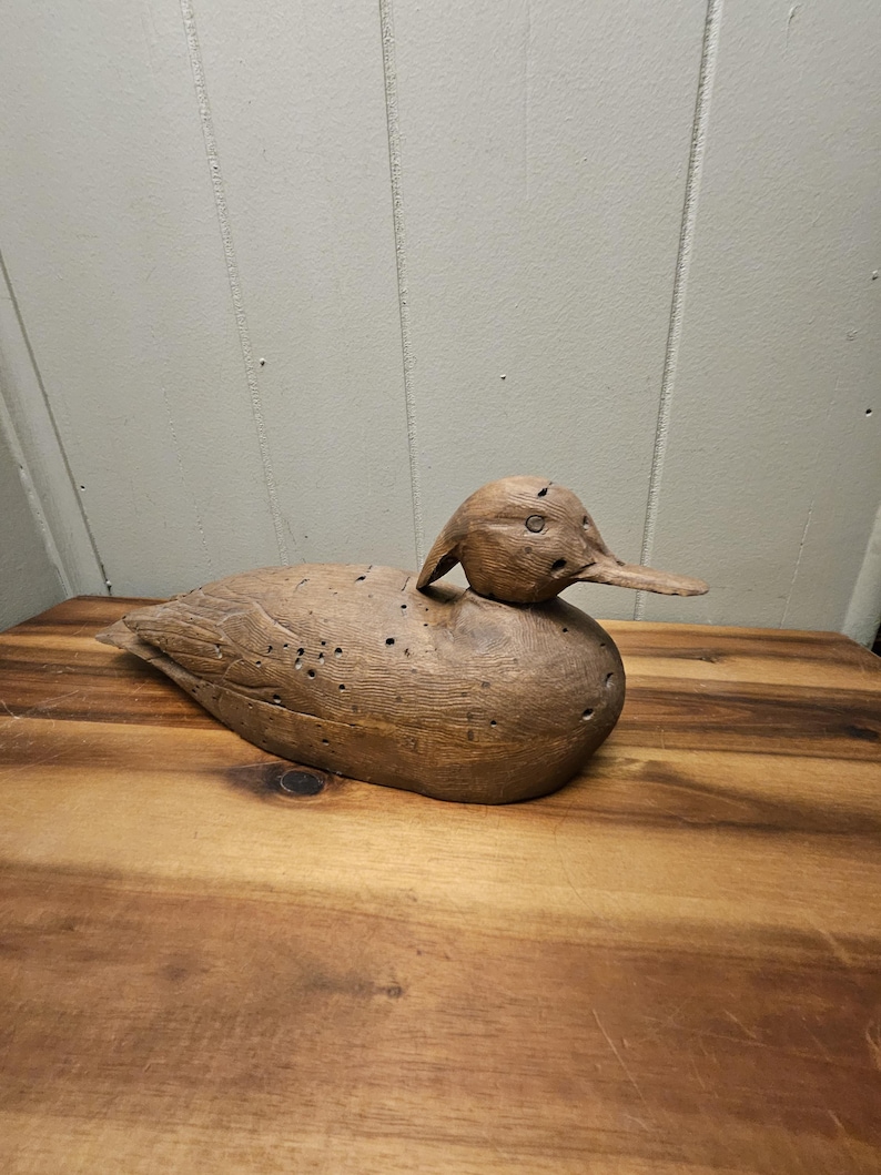 May include: A wooden duck decoy, carved from light brown wood. The duck has a detailed head, beak, and body, with small holes throughout. The decoy is placed on a wooden surface.