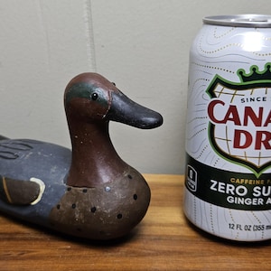 May include: A wooden duck decoy with brown, black, and teal paint, next to a 12 fl oz (355 ml) can of Canada Dry Zero Sugar Ginger Ale. The can is white with red and green text.