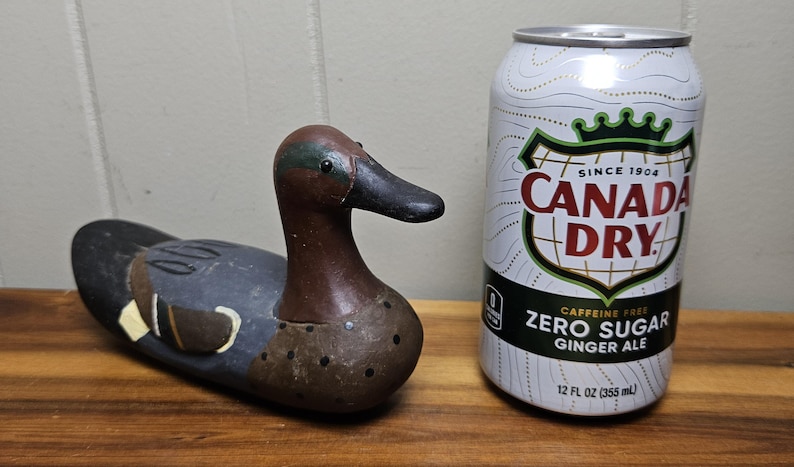 May include: A wooden duck decoy with brown, black, and teal paint, next to a 12 fl oz (355 ml) can of Canada Dry Zero Sugar Ginger Ale. The can is white with red and green text.