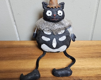 Puede incluir: Una figura de gato negro con cuerpo redondo, marcas esqueléticas pintadas en blanco y un sombrero puntiagudo de color tostado. El gato presenta un cuello con volantes plateados, brazos negros pequeños y largas piernas de cordón negro.