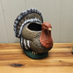 May include: A ceramic turkey ornament with a brown body, red head, and a fan of black, white, and brown feathers. The turkey sits on a green base and has a hollowed-out centre.