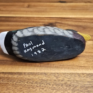 Puede incluir: Un se&ntilde;uelo de pato de madera pintado a mano, con una parte inferior negra y la inscripci&oacute;n "Paul Raymond 1982". El pato tiene una cabeza blanca, alas grises y negras y un pico amarillo. El se&ntilde;uelo descansa sobre una superficie de madera.