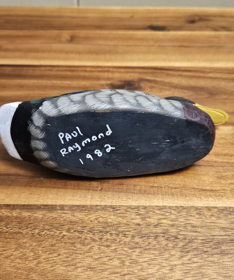 Puede incluir: Un se&ntilde;uelo de pato de madera pintado a mano, con una parte inferior negra y la inscripci&oacute;n "Paul Raymond 1982". El pato tiene una cabeza blanca, alas grises y negras y un pico amarillo. El se&ntilde;uelo descansa sobre una superficie de madera.