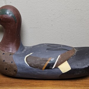 May include: A hand-painted wooden duck decoy. The duck features a brown head and neck, a black beak, and a body painted in shades of gray, brown, and white. The decoy is a decorative item.