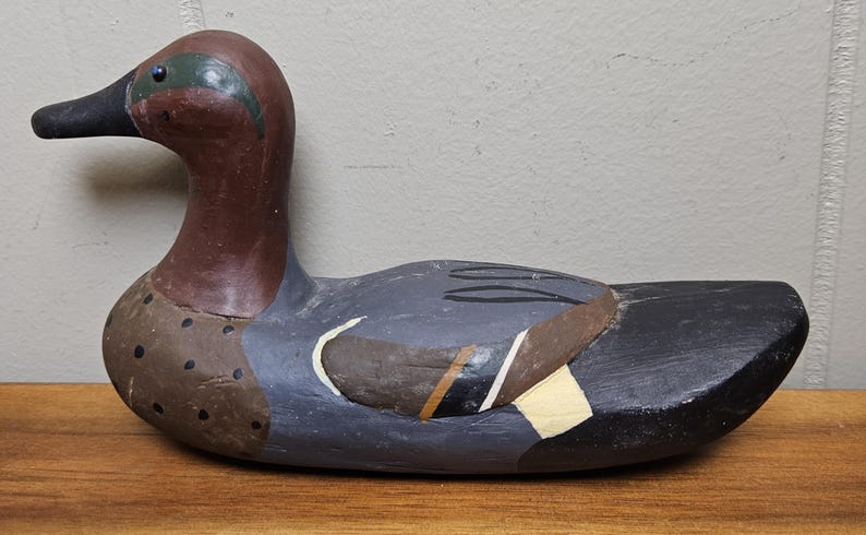 May include: A hand-painted wooden duck decoy. The duck features a brown head and neck, a black beak, and a body painted in shades of gray, brown, and white. The decoy is a decorative item.
