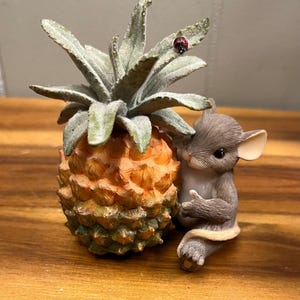 May include: A decorative figurine featuring a detailed pineapple with a green and gray leafy top. A small, gray mouse is nestled next to the pineapple. A red ladybug rests on one of the leaves. The figurine is on a wooden surface.