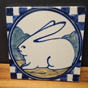 Op de afbeelding: Een vierkante keramische tegel met een handgeschilderd blauw-wit ontwerp. Een centrale cirkelvormige omlijsting bevat het silhouet van een wit konijn tegen een gedempt groen en beige landschap. De rand bestaat uit een blauw-wit geblokt patroon.
