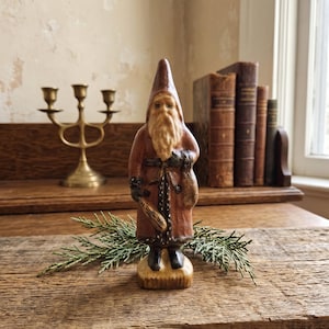 Könnte beinhalten: Eine handgeschnitzte Holzfigur eines bärtigen Wesens mit einer hohen spitzen Mütze und einem langen braunen Mantel mit dunklem Besatz. Die Figur hält einen kleinen Sack und ein Holzwerkzeug in den Händen und steht auf einem runden Sockel.