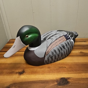 May include: A hand-painted wooden duck figurine. The duck has a green head, a white beak, and a body with gray, brown, and black feathers. The duck is resting on a wooden surface. The figurine is a decorative item.