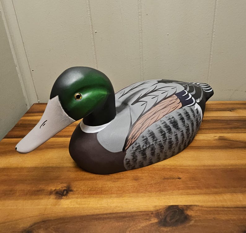 May include: A hand-painted wooden duck figurine. The duck has a green head, a white beak, and a body with gray, brown, and black feathers. The duck is resting on a wooden surface. The figurine is a decorative item.