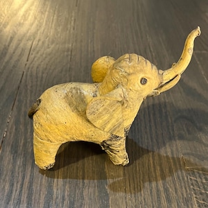 Può includere: Una piccola statuetta di elefante realizzata a mano in pelle marrone chiaro. L'elefante ha la proboscide alzata e dettagli elaborati.