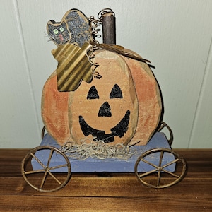 May include: A wooden pumpkin with a carved jack-o'-lantern face on wheels. The pumpkin is painted orange and has a black cat on top. The wheels are made of metal and are painted blue.