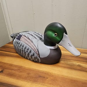 May include: A hand-painted wooden duck decoy. The duck has a green head, white beak, and a body with gray, brown, and black markings. The decoy is resting on a wooden surface. The duck is a decorative item.