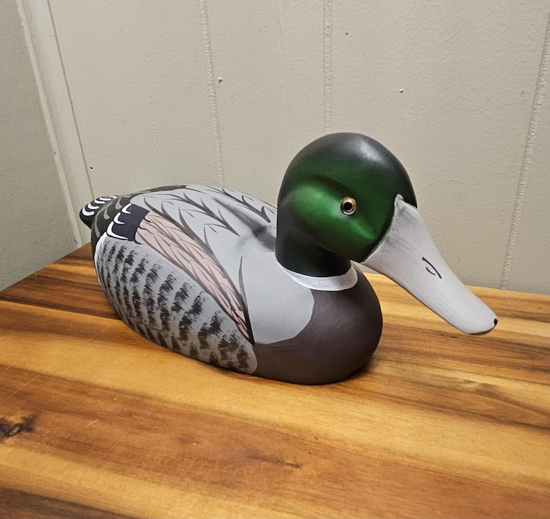 May include: A hand-painted wooden duck decoy. The duck has a green head, white beak, and a body with gray, brown, and black markings. The decoy is resting on a wooden surface. The duck is a decorative item.