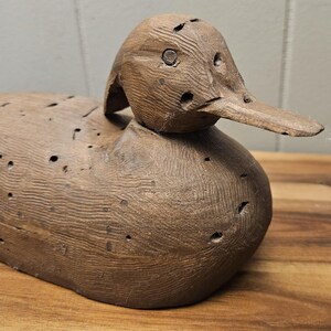 May include: A weathered, wooden duck decoy with a detailed, carved appearance. The duck has a long, flat beak and visible holes, suggesting it's an antique or vintage piece. The wood grain is prominent, and the color is a warm brown.