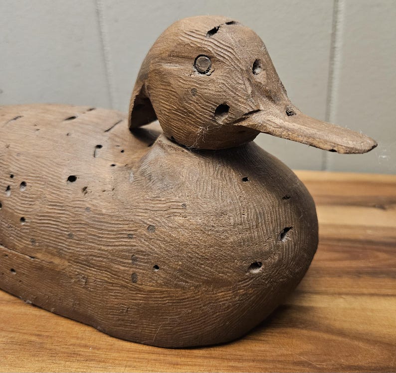 May include: A weathered, wooden duck decoy with a detailed, carved appearance. The duck has a long, flat beak and visible holes, suggesting it's an antique or vintage piece. The wood grain is prominent, and the color is a warm brown.