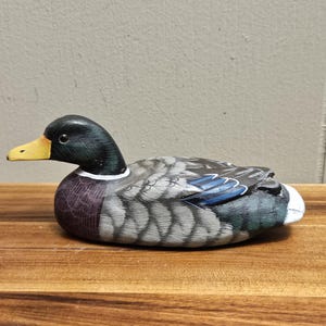Puede incluir: Figura de pato de madera pintada a mano. El pato tiene una cabeza negra con pico amarillo, cuerpo con dise&ntilde;o gris y azul y cola blanca. El pato est&aacute; sobre una superficie de madera.