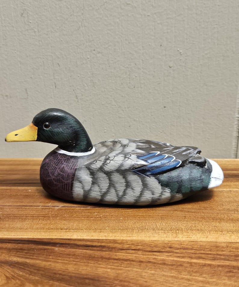 Puede incluir: Figura de pato de madera pintada a mano. El pato tiene una cabeza negra con pico amarillo, cuerpo con dise&ntilde;o gris y azul y cola blanca. El pato est&aacute; sobre una superficie de madera.