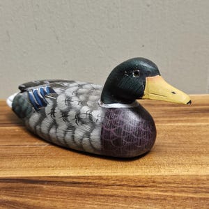 Puede incluir: Figura de pato de madera pintada a mano. El pato tiene una cabeza negra, un pico amarillo y un cuerpo con plumas grises, azules y moradas. La figura est&aacute; sobre una superficie de madera.