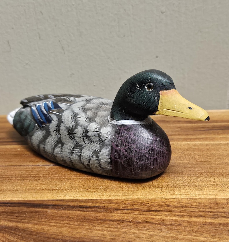 Puede incluir: Figura de pato de madera pintada a mano. El pato tiene una cabeza negra, un pico amarillo y un cuerpo con plumas grises, azules y moradas. La figura est&aacute; sobre una superficie de madera.