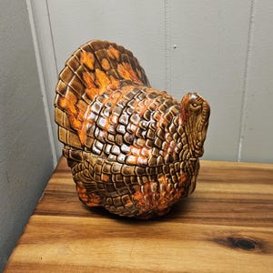 May include: A ceramic turkey figurine with a detailed, textured design. The turkey features shades of brown, orange, and tan, with a fanned tail. The figurine is likely a decorative item, possibly for Thanksgiving or fall decor.