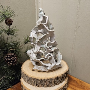 May include: A white ceramic sculpture with an abstract, organic design. The sculpture is placed on a wooden base made of two stacked tree slices. The piece has a textured surface and a unique, artistic aesthetic.