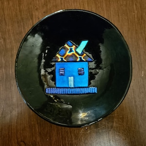 May include: A black ceramic dish with a blue and yellow house design. The house has a blue roof and a yellow and black patterned front.