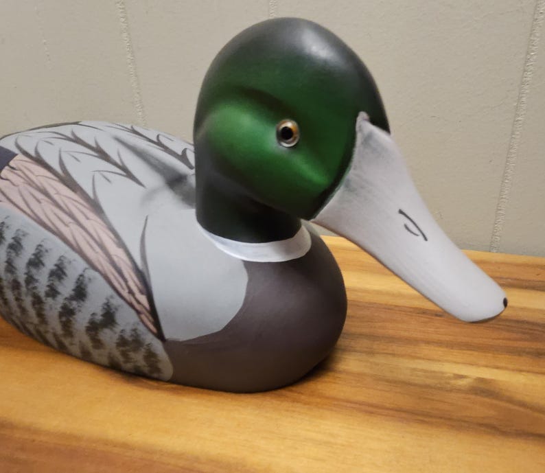 May include: A hand-painted wooden duck decoy. The duck has a green head, white beak, and a white collar. The body is painted in shades of gray, brown, and white, with detailed feather patterns. The decoy is resting on a wooden surface.