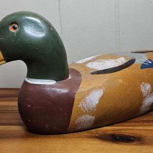 K&ouml;nnte beinhalten: Ein h&ouml;lzerner Entenk&ouml;der mit gr&uuml;nem Kopf, gelbem Schnabel und braunem K&ouml;rper mit wei&szlig;en und schwarzen Akzenten. Die Ente liegt auf einer Holzoberfl&auml;che und zeigt ihre detaillierte Handwerkskunst und ihren rustikalen Charme.