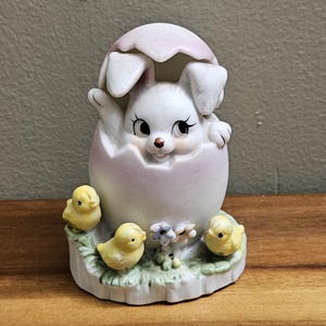 May include: A white ceramic bunny figurine peeking out of a pink and white egg with three yellow chicks surrounding the base. The figurine is decorated with flowers and green leaves.