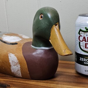 K&ouml;nnte beinhalten: Eine h&ouml;lzerne Entenattrappe mit gr&uuml;nem Kopf, gelbem Schnabel und braunem K&ouml;rper, neben einer Dose Canada Dry Zero Sugar Ginger Ale. Die Dose ist wei&szlig; mit einem gr&uuml;nen und schwarzen Logo und dem Text "CANADA DRY" und "ZERO SUGAR GINGER ALE".