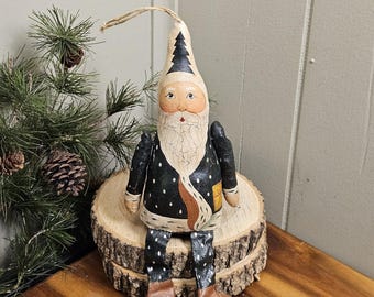 May include: A handmade Santa Claus figure with a long, pointed hat decorated with a black Christmas tree design. The Santa figure wears a black coat with white polka dots and brown boots. It is seated on a wooden slice, with a small evergreen tree in the background.