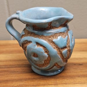 May include: A small, light blue ceramic pitcher with a handle and spout. The pitcher features a textured, brown band around the middle with a swirling design. The pitcher is sitting on a wooden surface.
