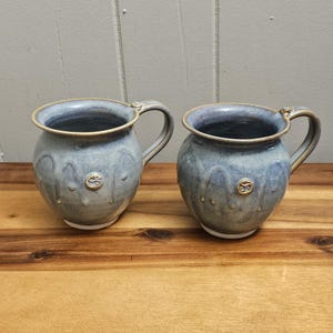 Może przedstawiać: Dwa niebieskie ceramiczne kubki z fakturowanym szkliwem i złotymi akcentami. Kubki mają zaokrąglony kształt i uchwyt po boku.