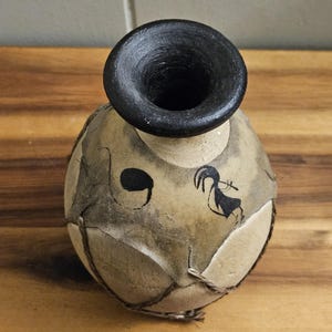 Puede incluir: Un jarrón esférico artesanal con borde negro y cuerpo beige. El jarrón presenta un diseño pintado en negro de una persona tocando la flauta. El jarrón está decorado con una cuerda trenzada.