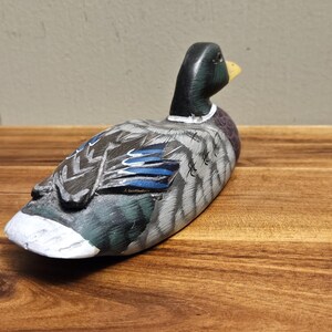 Puede incluir: Un se&ntilde;uelo de pato de madera pintado a mano. El pato presenta una cabeza negra, un pico amarillo y un cuerpo con plumas grises, azules y blancas. El se&ntilde;uelo est&aacute; colocado sobre una superficie de madera, mostrando la artesan&iacute;a detallada y la apariencia realista.