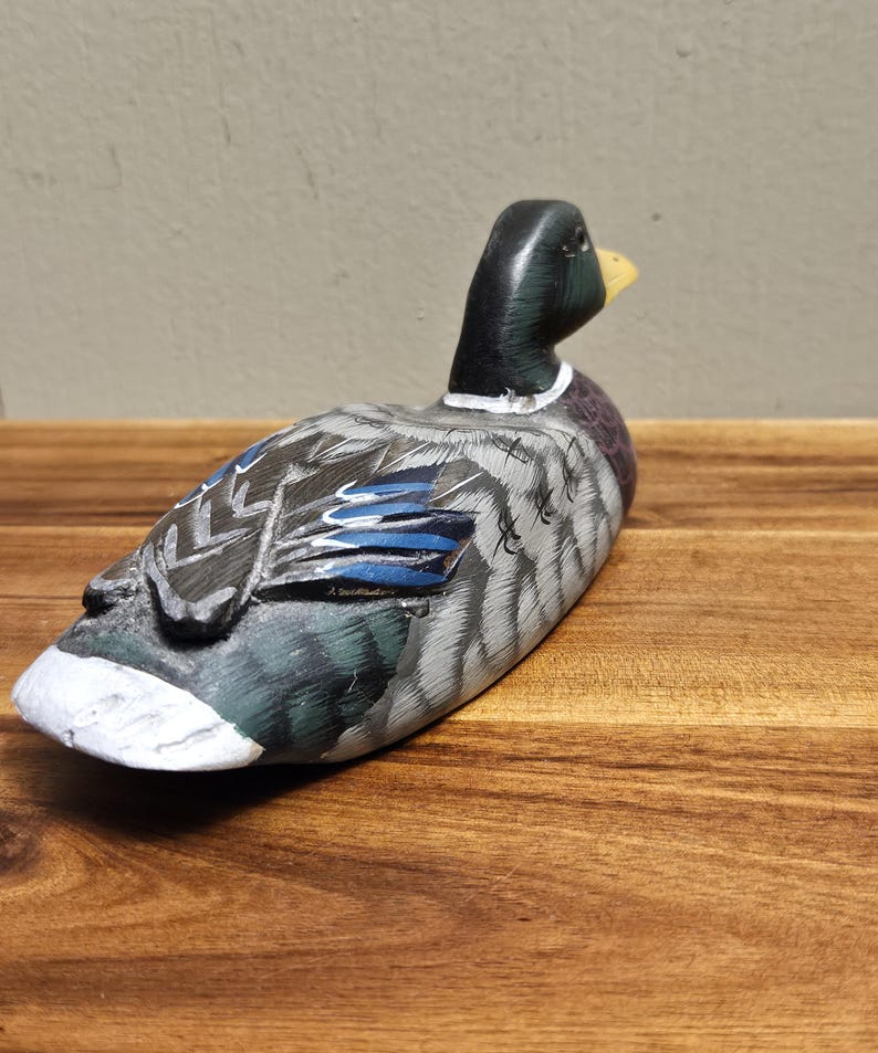 Puede incluir: Un se&ntilde;uelo de pato de madera pintado a mano. El pato presenta una cabeza negra, un pico amarillo y un cuerpo con plumas grises, azules y blancas. El se&ntilde;uelo est&aacute; colocado sobre una superficie de madera, mostrando la artesan&iacute;a detallada y la apariencia realista.