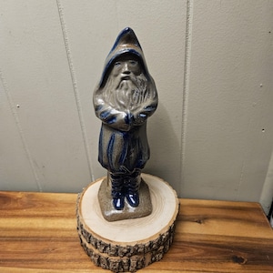 Puede incluir: Figura de cerámica de un mago con una túnica azul con capucha, con una larga barba blanca y los brazos cruzados. El mago está sobre una pequeña base cuadrada, encima de una rodaja de madera con bordes de corteza. Aspecto artesanal.