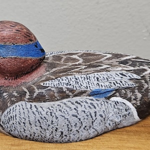Op de afbeelding: Een handgesneden houten eendenbeeldje. De eend heeft een bruine kop met een blauwe streep, een zwarte snavel en gedetailleerde veermotieven in bruin-, grijs- en wittinten. De eend rust op een houten oppervlak.