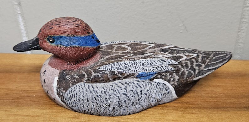 Op de afbeelding: Een handgesneden houten eendenbeeldje. De eend heeft een bruine kop met een blauwe streep, een zwarte snavel en gedetailleerde veermotieven in bruin-, grijs- en wittinten. De eend rust op een houten oppervlak.