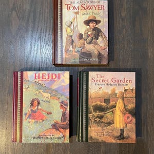 Op de afbeelding: Drie vintage hardcover boeken met geïllustreerde omslagen. Het bovenste boek is "De avonturen van Tom Sawyer" van Mark Twain, het middelste boek is "Heidi" van Johanna Spyri, en het onderste boek is "De geheime tuin" van Frances Hodgson Burnett.