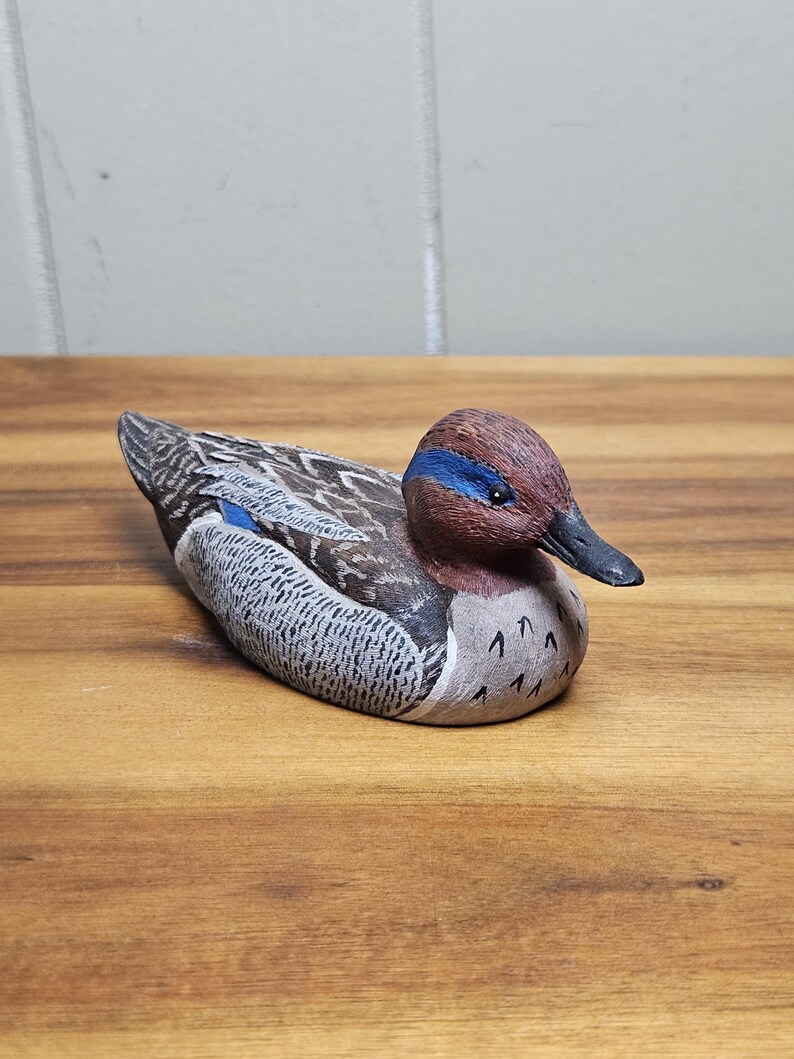 Op de afbeelding: Een handgesneden houten eendenbeeldje. De eend heeft een bruine kop met een blauwe streep, een zwarte snavel en een lichaam met grijze, bruine en witte details. De eend rust op een houten oppervlak.