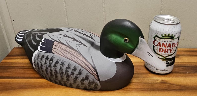 May include: A hand-painted wooden duck decoy with a green head, white beak, and detailed gray and brown feathers. A can of Canada Dry ginger ale is next to the duck. The can is white with red and black text.