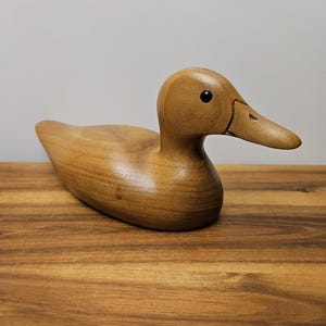 May include: A wooden duck figurine with a smooth, polished finish. The duck is light brown with a dark eye and a detailed beak. The figurine is sitting on a wooden surface.