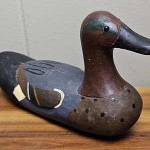 May include: A wooden duck decoy with a brown head and a black beak. The body is painted in shades of gray, brown, and black, with accents of yellow and white. The decoy is resting on a wooden surface.