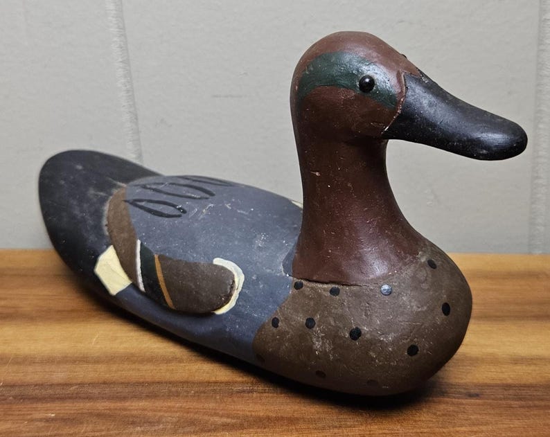 May include: A wooden duck decoy with a brown head and a black beak. The body is painted in shades of gray, brown, and black, with accents of yellow and white. The decoy is resting on a wooden surface.