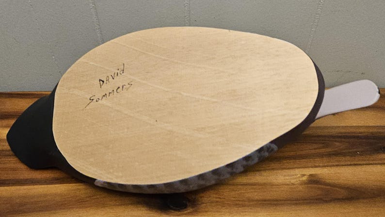 May include: A wooden duck decoy with a light brown oval-shaped top and a black body. The text "David Sommers" is handwritten on the top. The decoy has a gray tail.