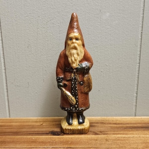 May include: A vintage Santa Claus figurine with a red and brown robe, white beard, and black boots. He is holding a sack and a stick in his hands. The figurine is standing on a small base.
