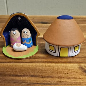 以下が含まれることがあります： 星でいっぱいの青と金の屋根、3体のフィギュア、緑色の土台が付いたセラミック製のクリスマスの飾り。隣には、青い屋根と黄色い窓が付いた小さな茶色のセラミック製の小屋がある。