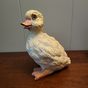 Puede incluir: Una pequeña figura de pato amarillo con una superficie texturizada. El pato tiene un ojo negro y un pico ligeramente abierto.
